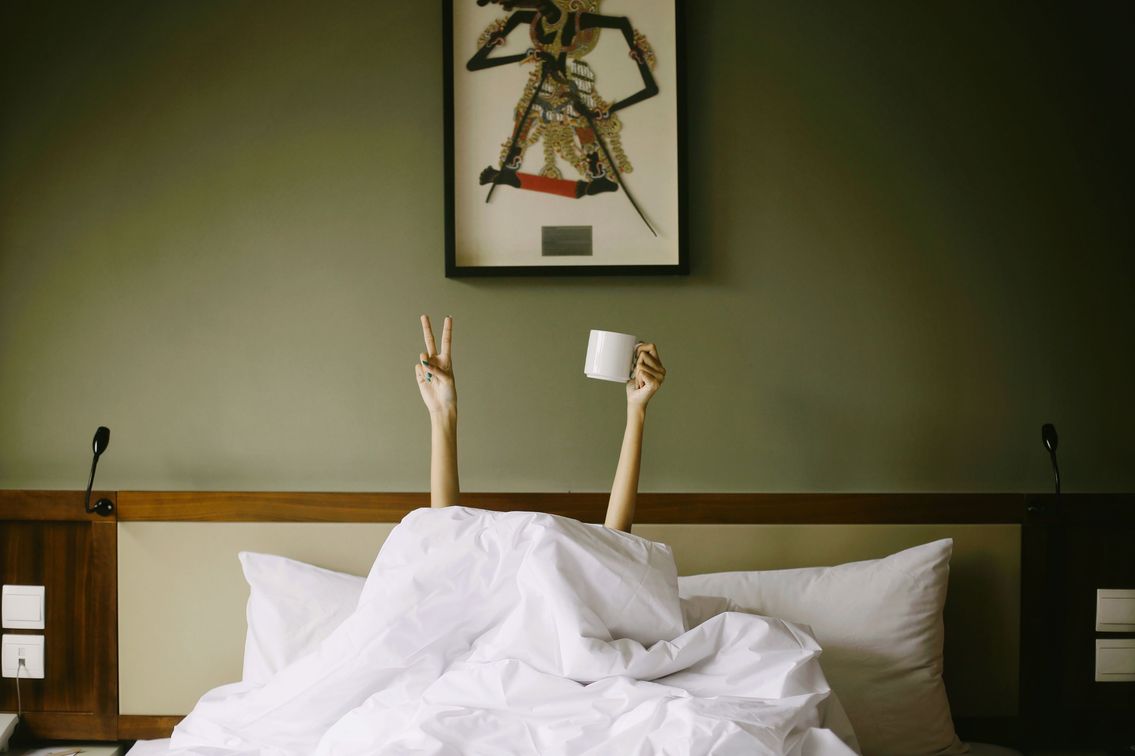 A person lying in bed with their feet up, holding a cup and making a peace sign.