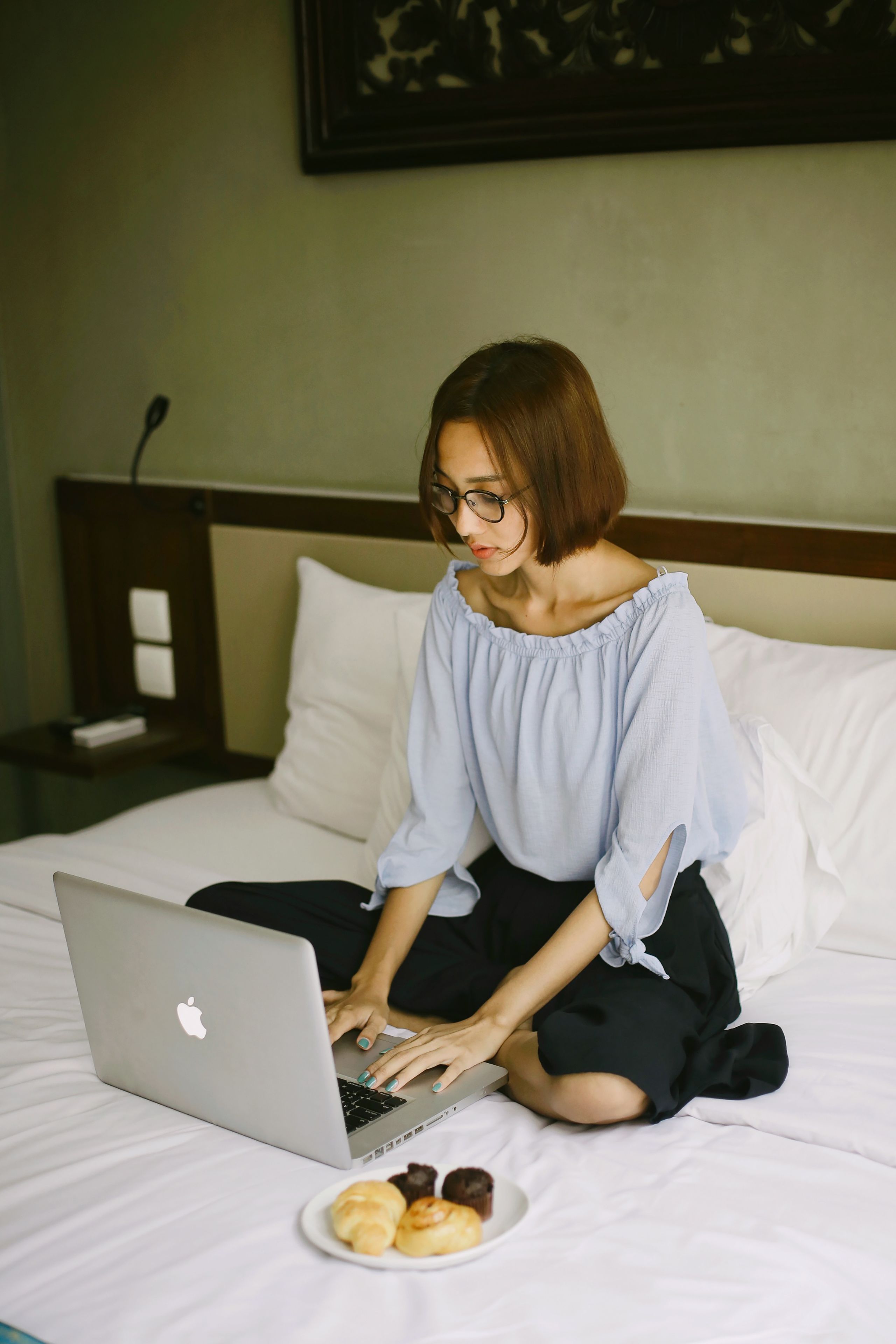A person sitting on a bed with a laptop, pillows, and a plate of pastries.