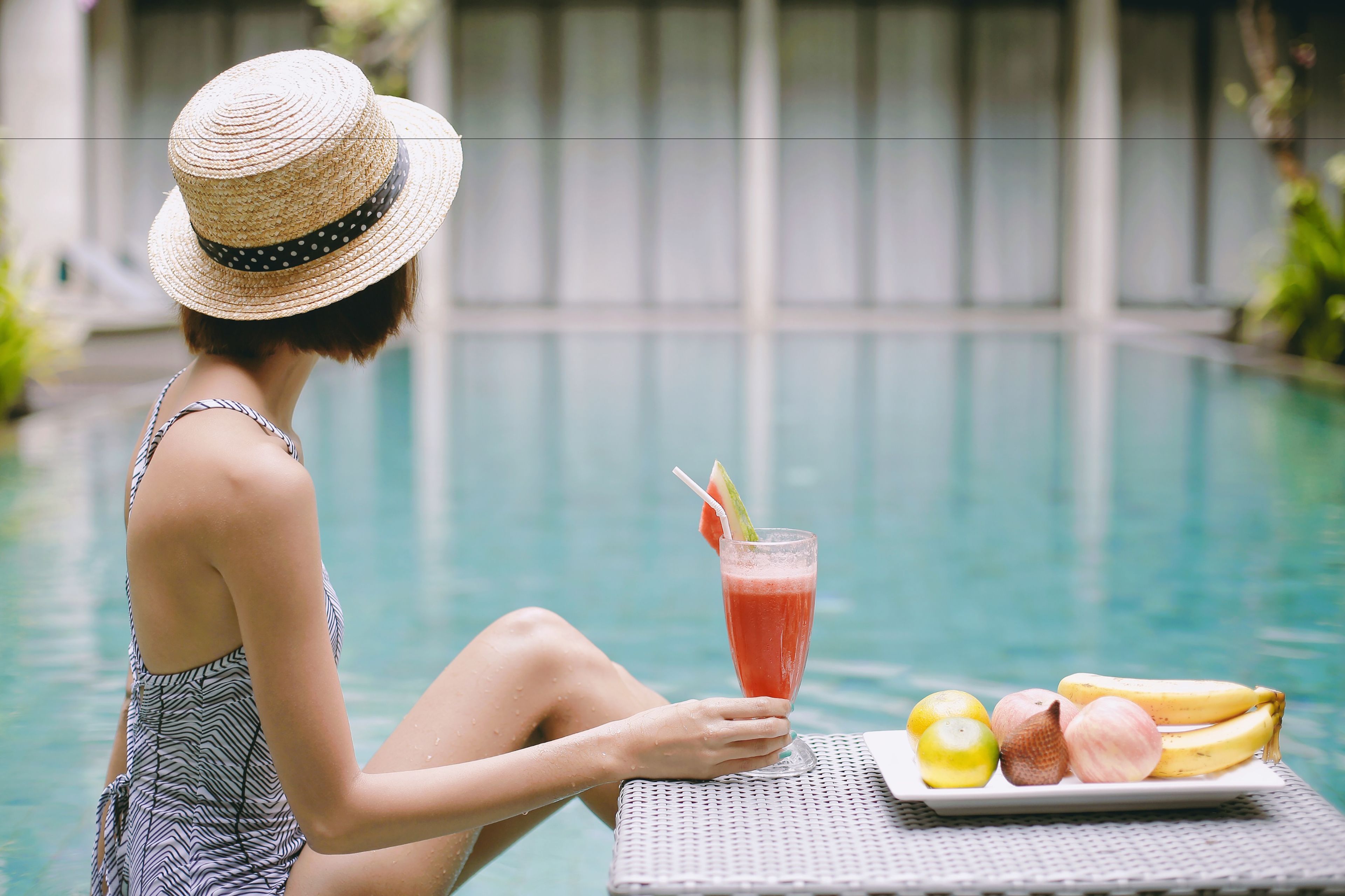 A person relaxing by a pool with a drink and fruit on a table.