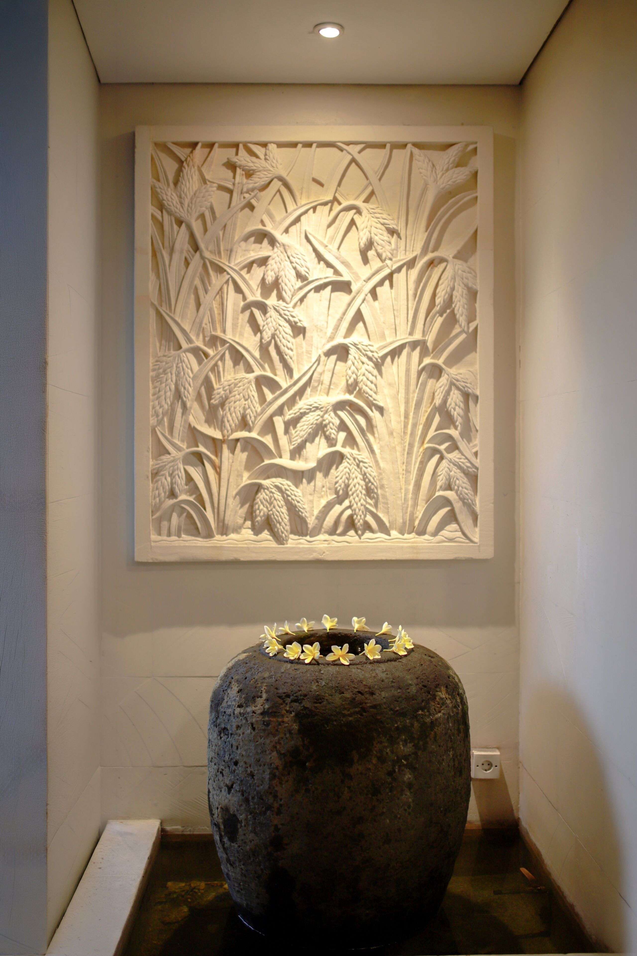 A wall with a relief sculpture of wheat and flowers, a large stone planter with flowers, and a recessed light.