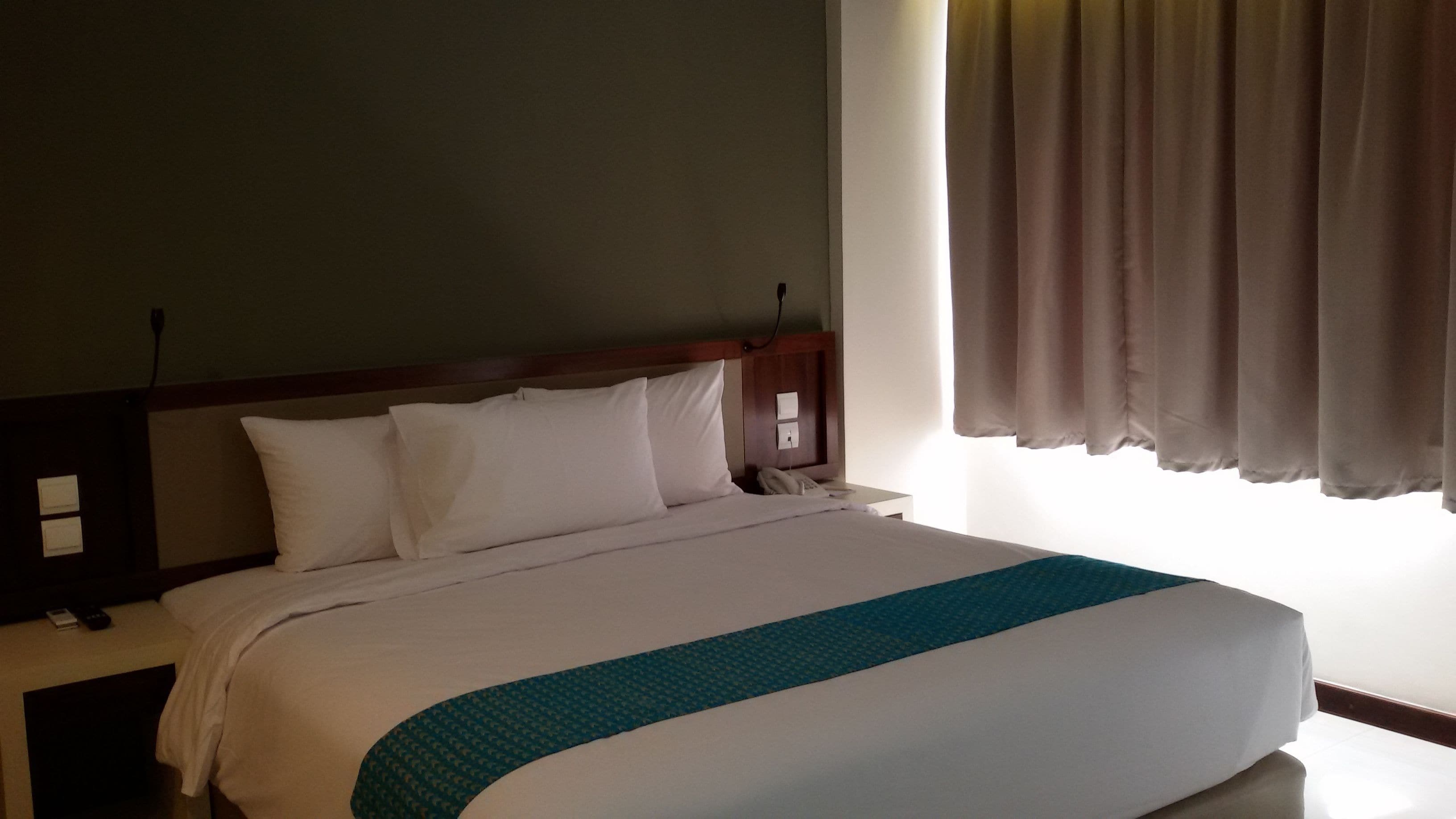 Habitación de hotel con cama, mesita de noche, lámpara, ventana con cortinas e interruptor de pared.