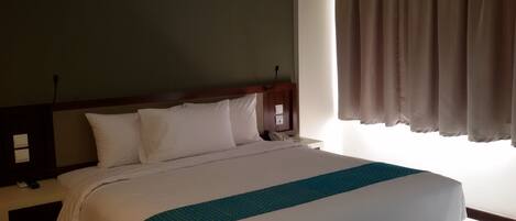Habitación de hotel con cama, mesita de noche, lámpara, ventana con cortinas e interruptor de pared.