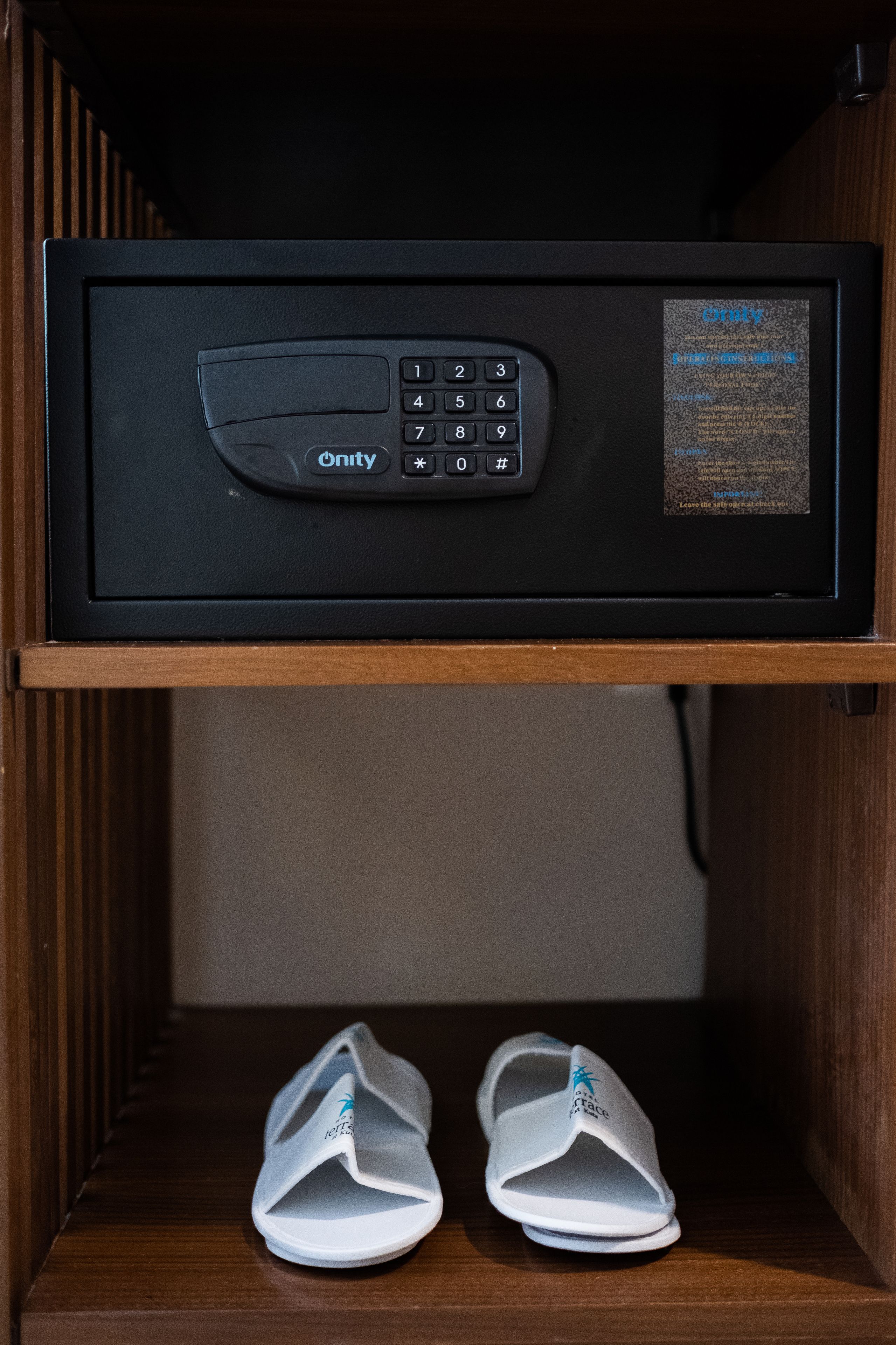 A microwave with a keypad and a label on the right side, placed above a wooden shelf with a pair of white slippers.