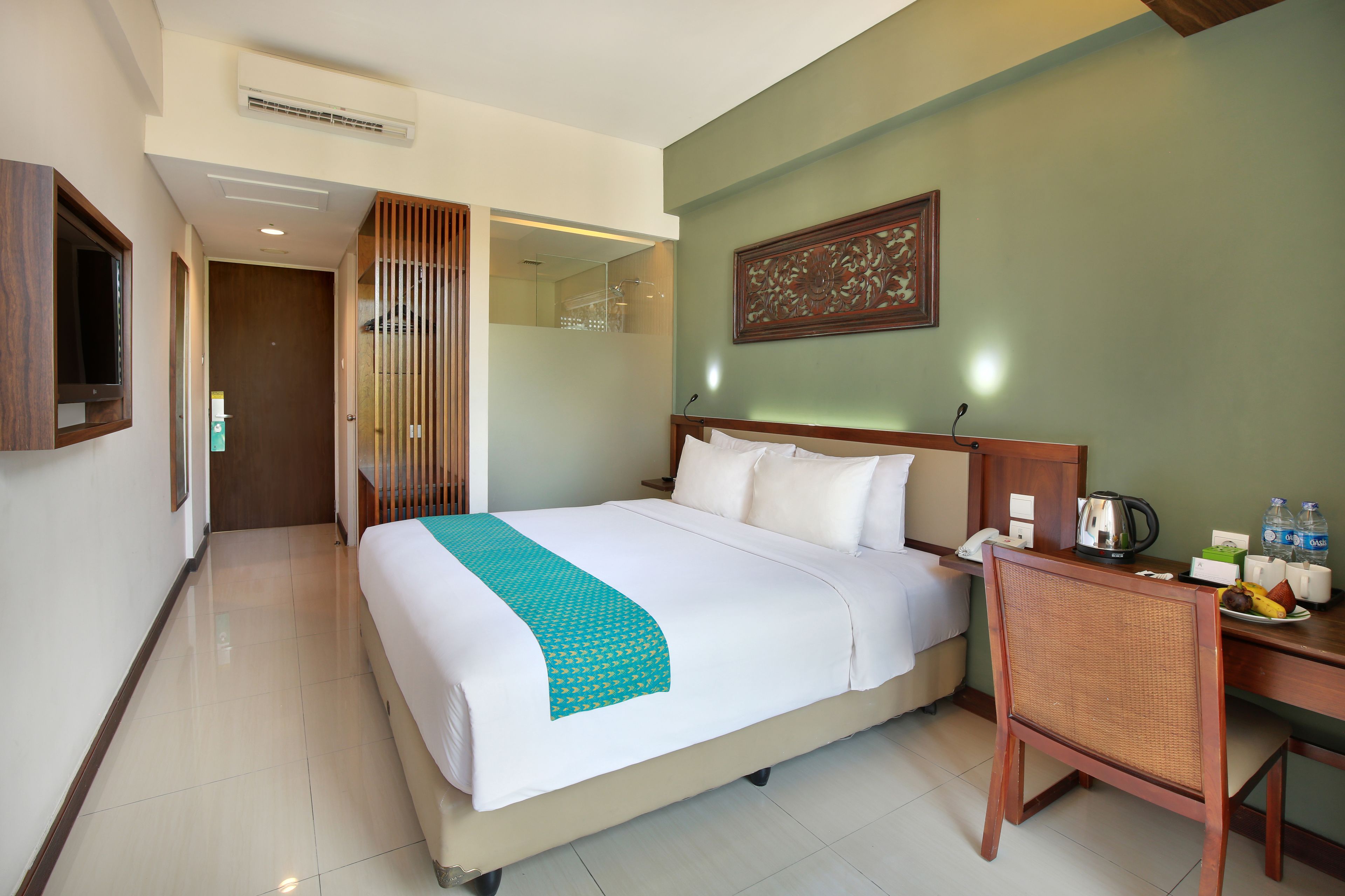 A hotel room with a bed, a desk, a chair, a mirror, a wall-mounted TV, and a door leading to another room.