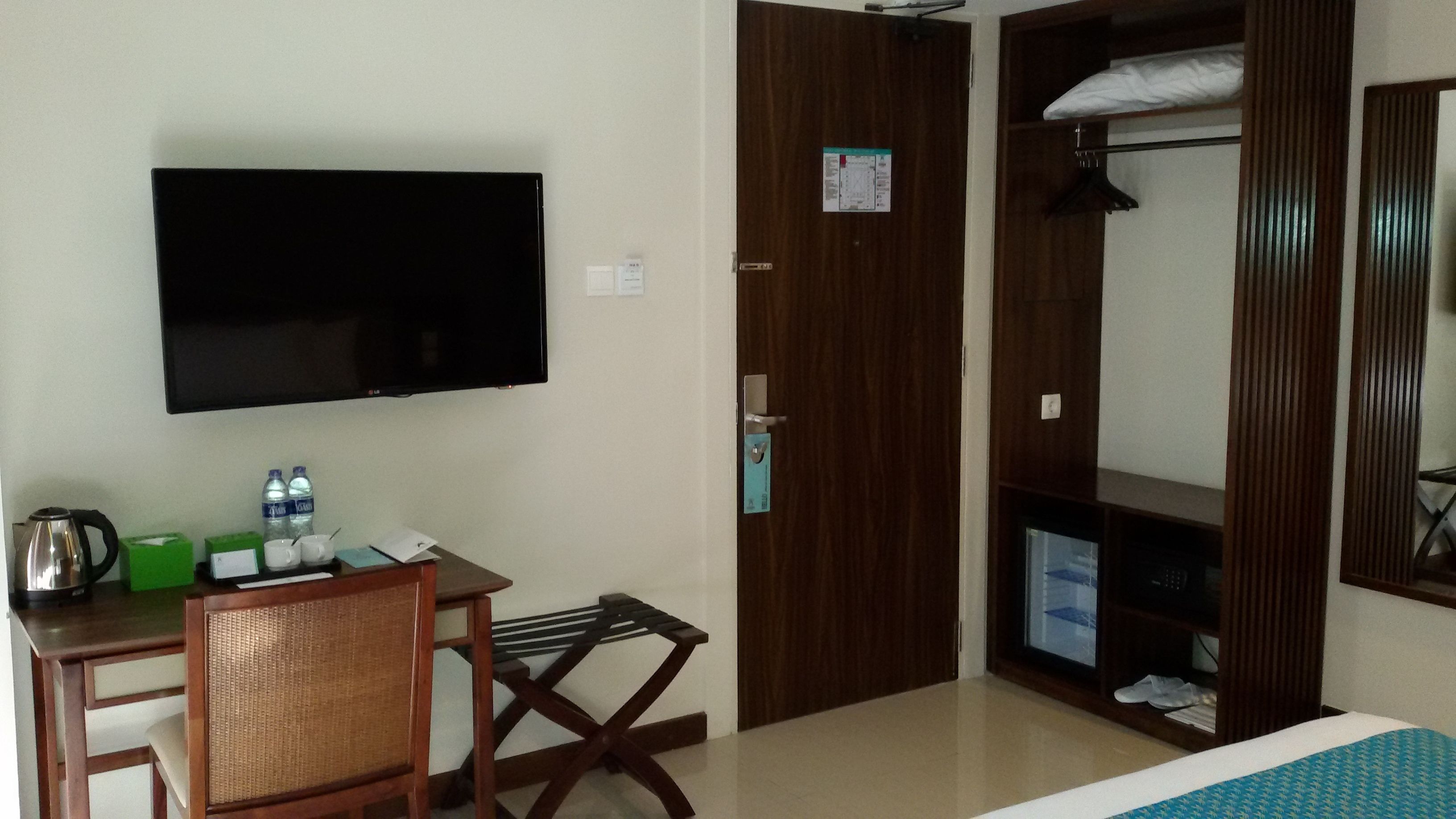 Habitación de hotel con cama, escritorio con silla, televisor, armario y una pequeña mesa con hervidor y botellas de agua.