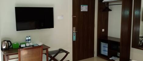 Habitación de hotel con cama, escritorio con silla, televisor, armario y una pequeña mesa con hervidor y botellas de agua.