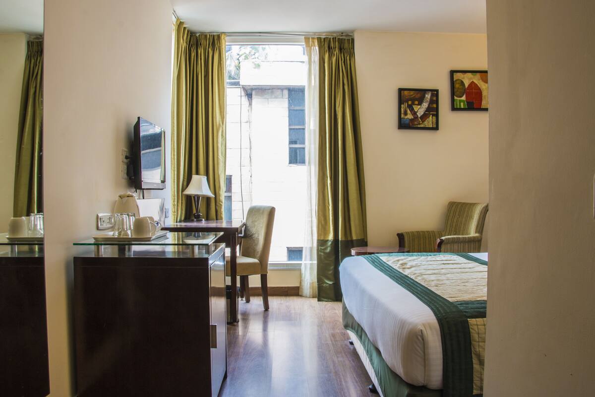 A hotel room with a bed, a desk with a lamp, a chair, a television, and a window with curtains.