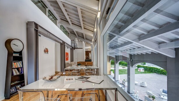 A modern interior with a long dining table, clear chairs, a bookshelf, and a view of the outside.
