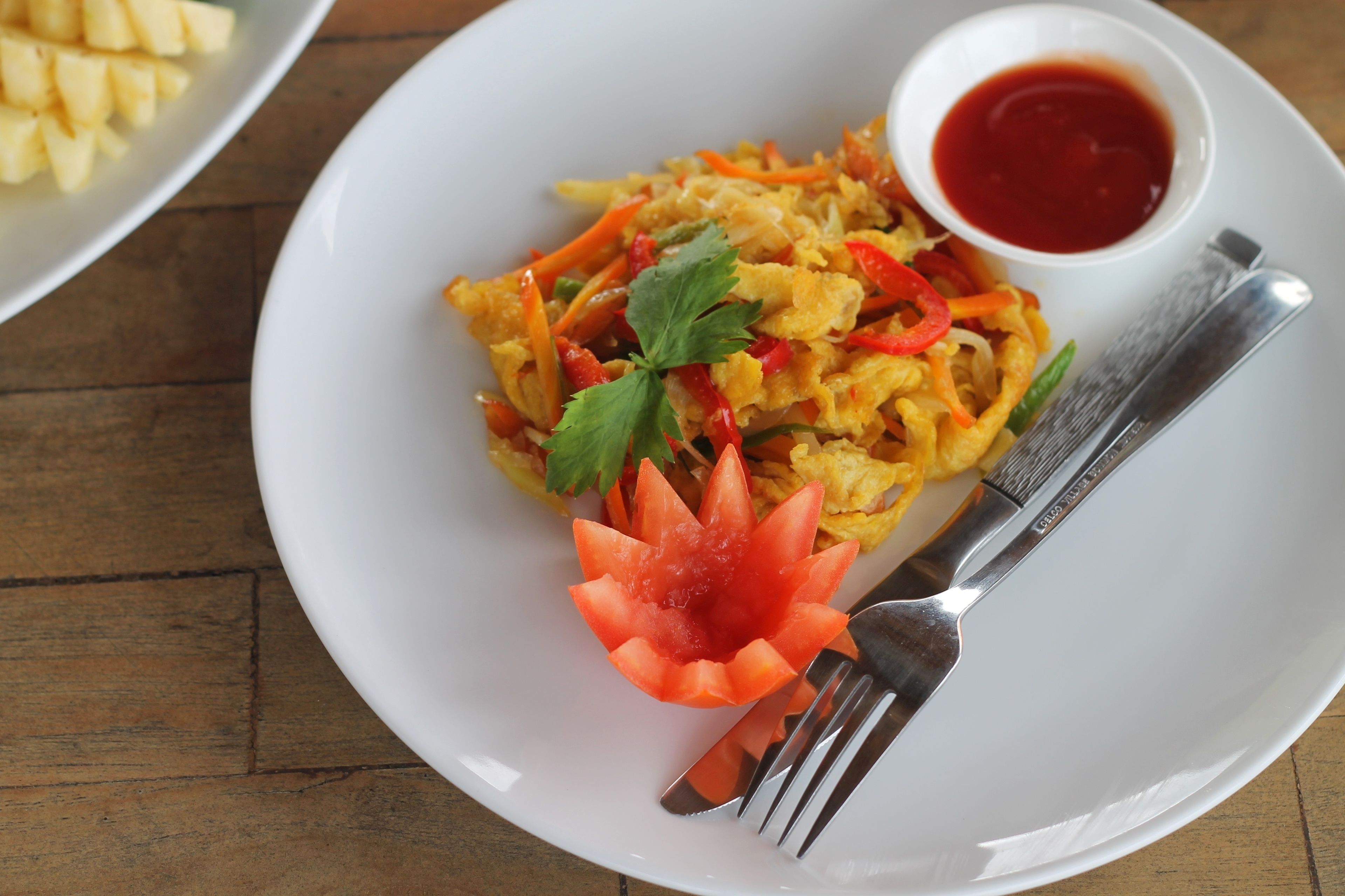 A white plate with a dish of stir-fried vegetables and a small bowl of red sauce, accompanied by a fork and knife on a wooden table.