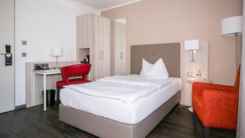 A hotel room with a bed, a desk, a chair, a lamp, and a trash bin.