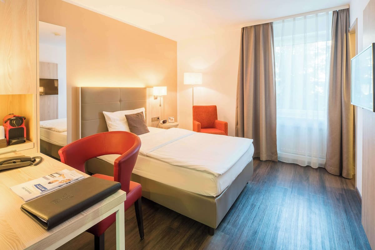 A hotel room with a bed, a desk with a chair, a red chair, a lamp, and a window with curtains.