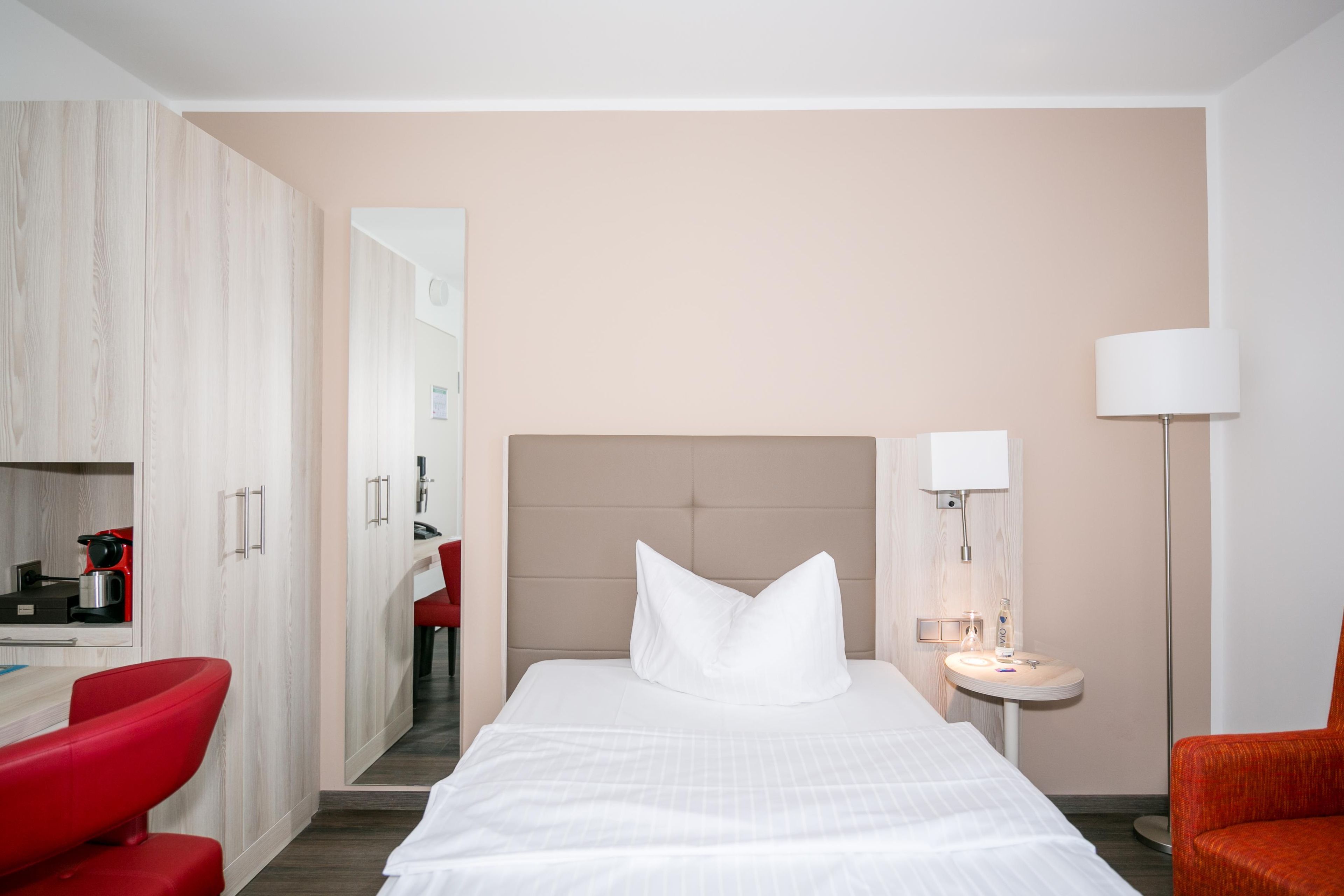 A hotel room with a bed, a red chair, a desk with a coffee machine, a mirror, and a bedside table with a lamp.