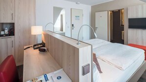 Une chambre d’hôtel équipée d’un lit, d’un bureau avec une lampe, d’un téléphone, d’un miroir et d’un téléviseur fixé au mur.