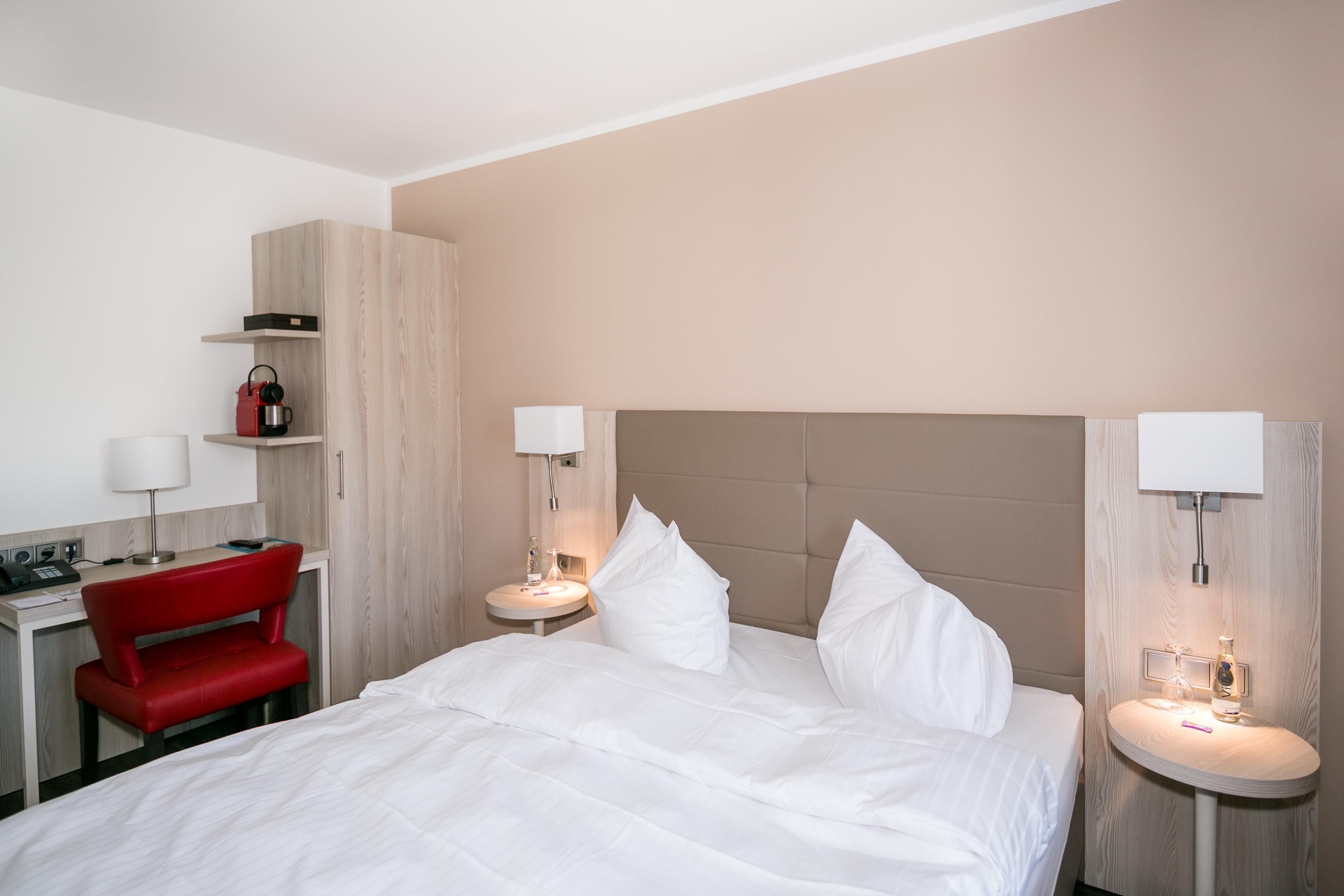Habitación de hotel con una cama, un escritorio, una silla roja y lámparas de noche.