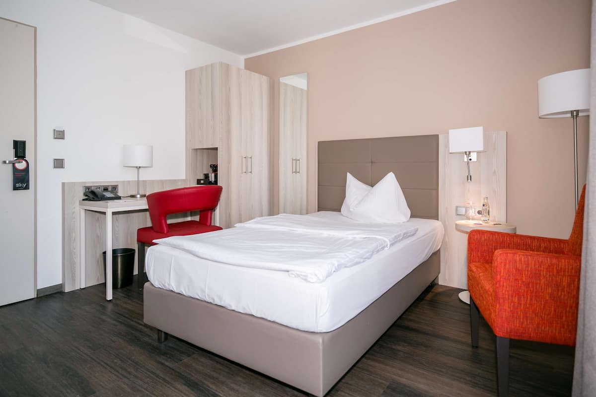 A hotel room with a bed, a desk, a chair, a lamp, and a trash bin.