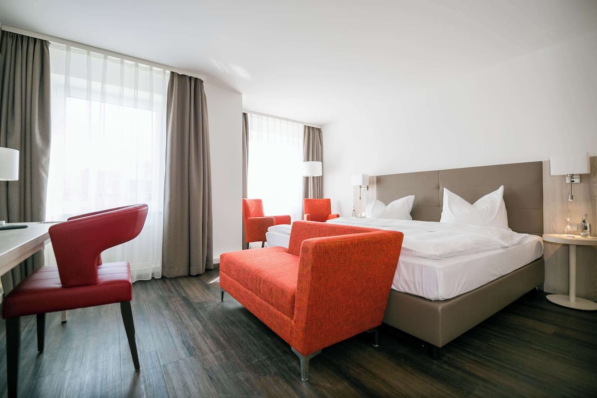 A hotel room with a bed, two red chairs, a desk, and a lamp.