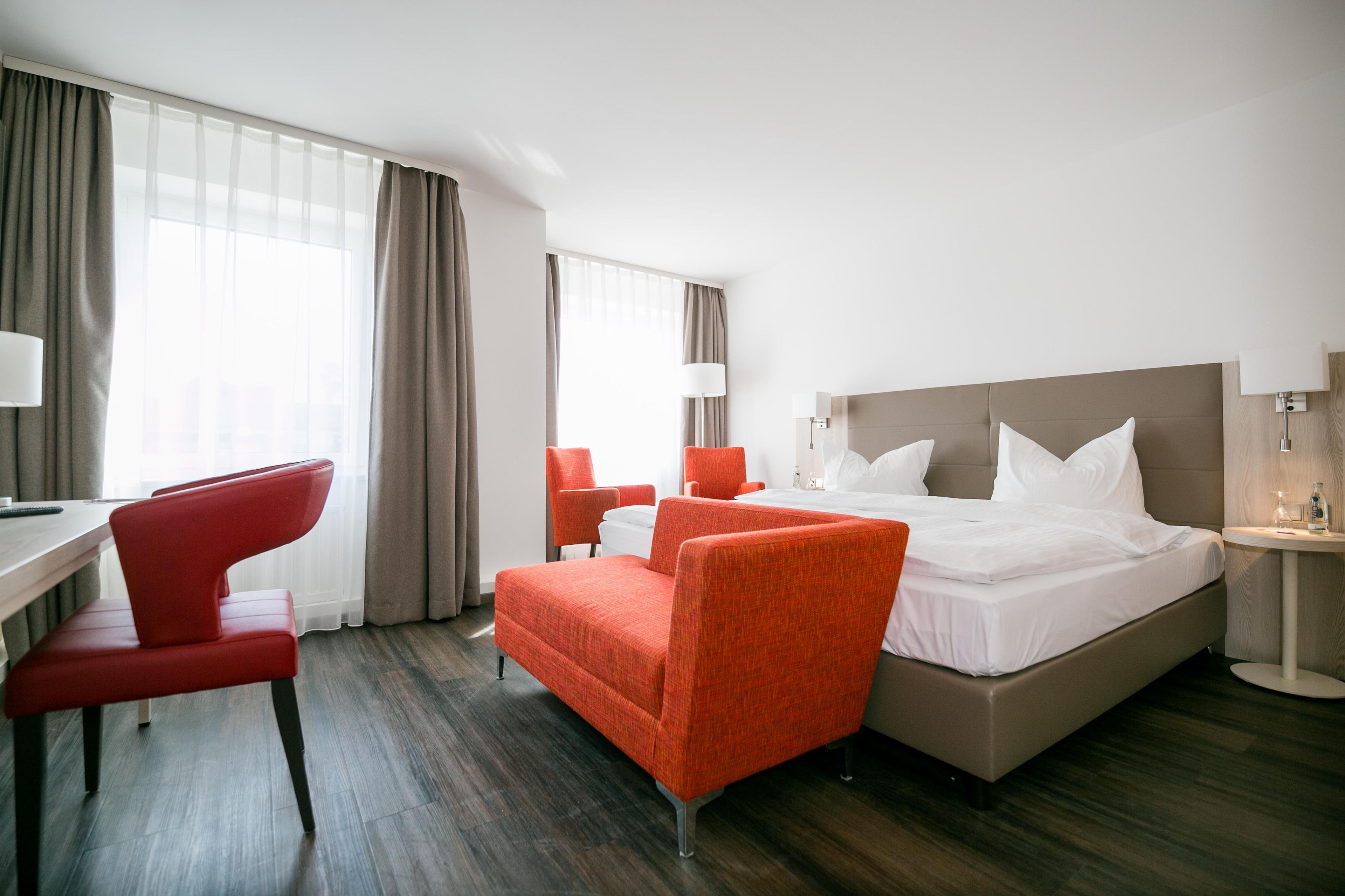 Una habitación de hotel con una cama, dos sillas rojas, un escritorio y una lámpara.