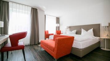 Una habitación de hotel con una cama, dos sillas rojas, un escritorio y una lámpara.