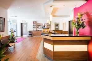 Zona de recepción de hotel con mostrador de madera, un área de descanso con sillas, un mostrador con computadora y un espejo grande que refleja la sala.