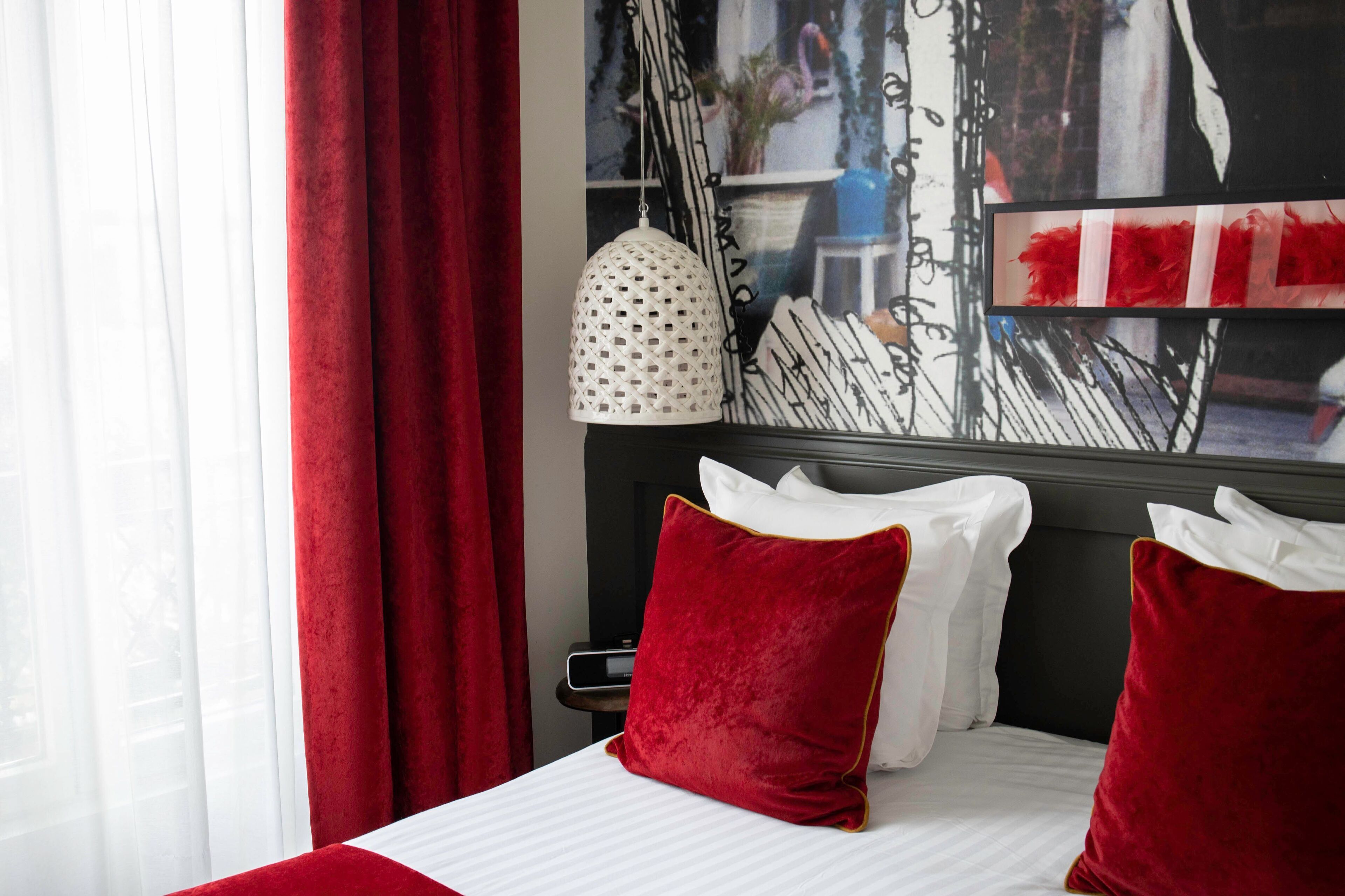 Ett hotellrum med en säng, röda och vita kuddar, en vit lampa och ett fönster med röda gardiner.