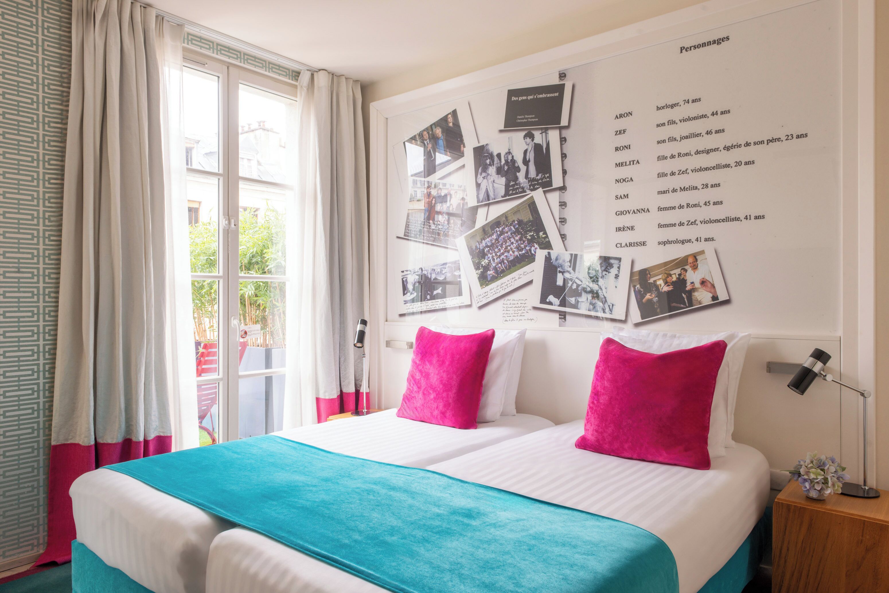 A hotel room with a bed, a wall with photos and names, a window with curtains, and a nightstand with a lamp.