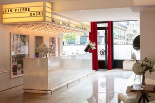 A hotel lobby with a reception desk, framed pictures on the walls, and a view of the street outside.
