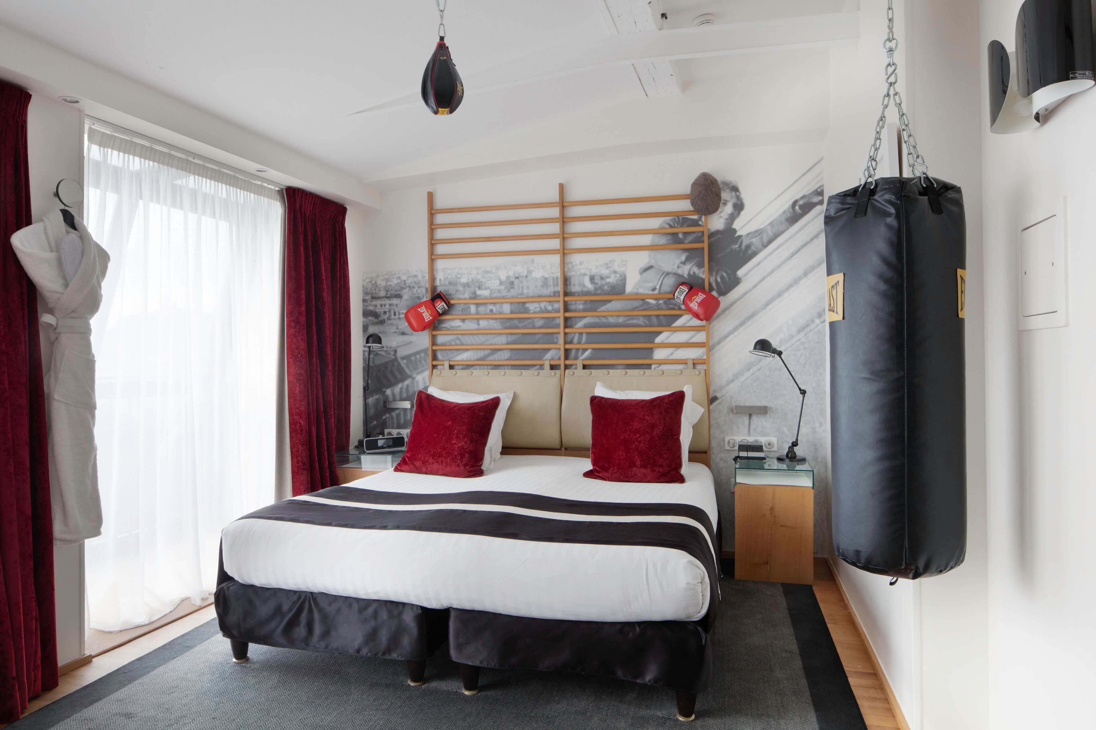 Habitación de hotel con cama, cortinas rojas, cabecera de madera, saco de boxeo y un mural con vistas urbanas en la pared.
