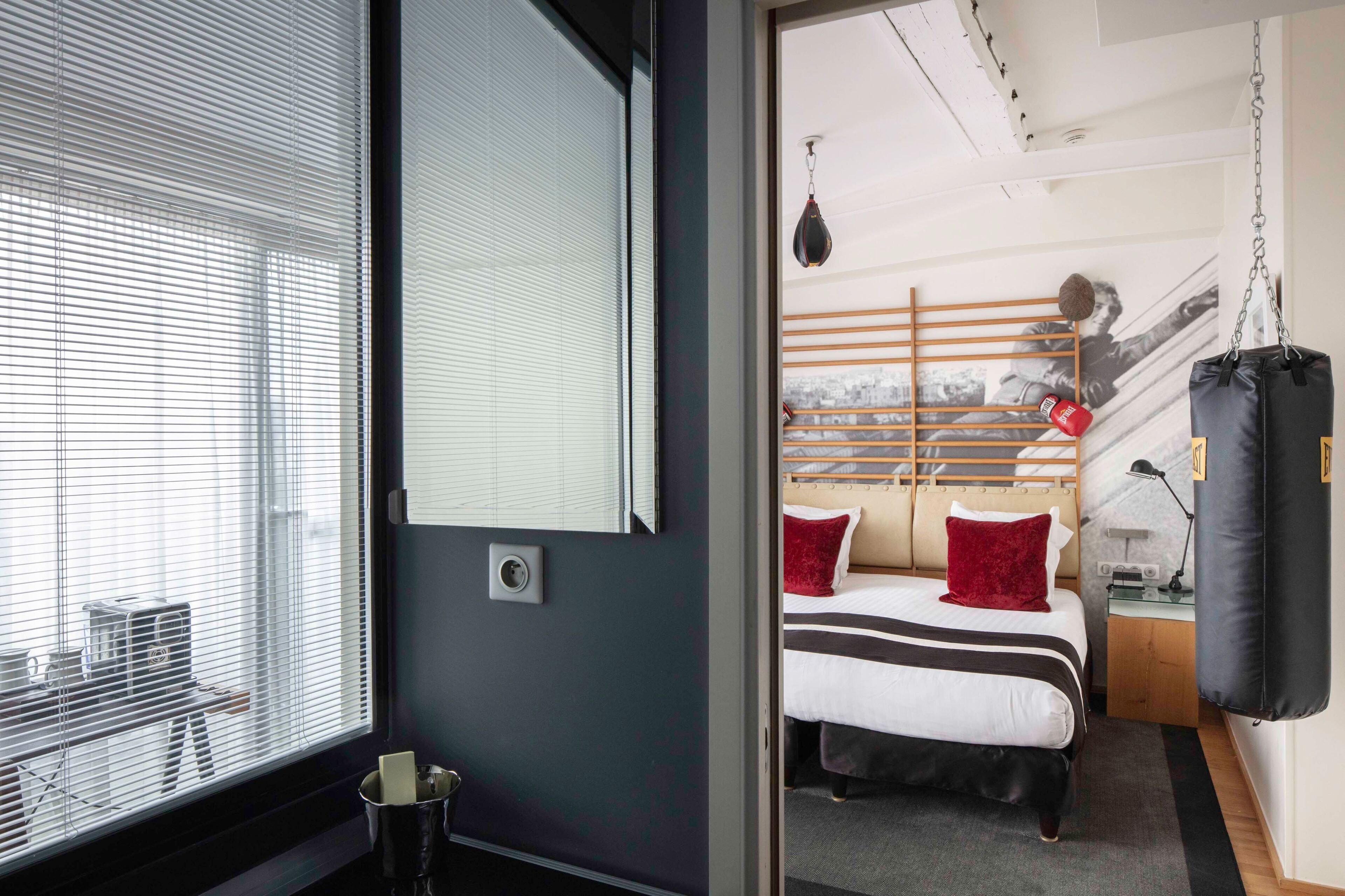 A hotel room with a bed, a punching bag, a window with blinds, and a view of the outside.