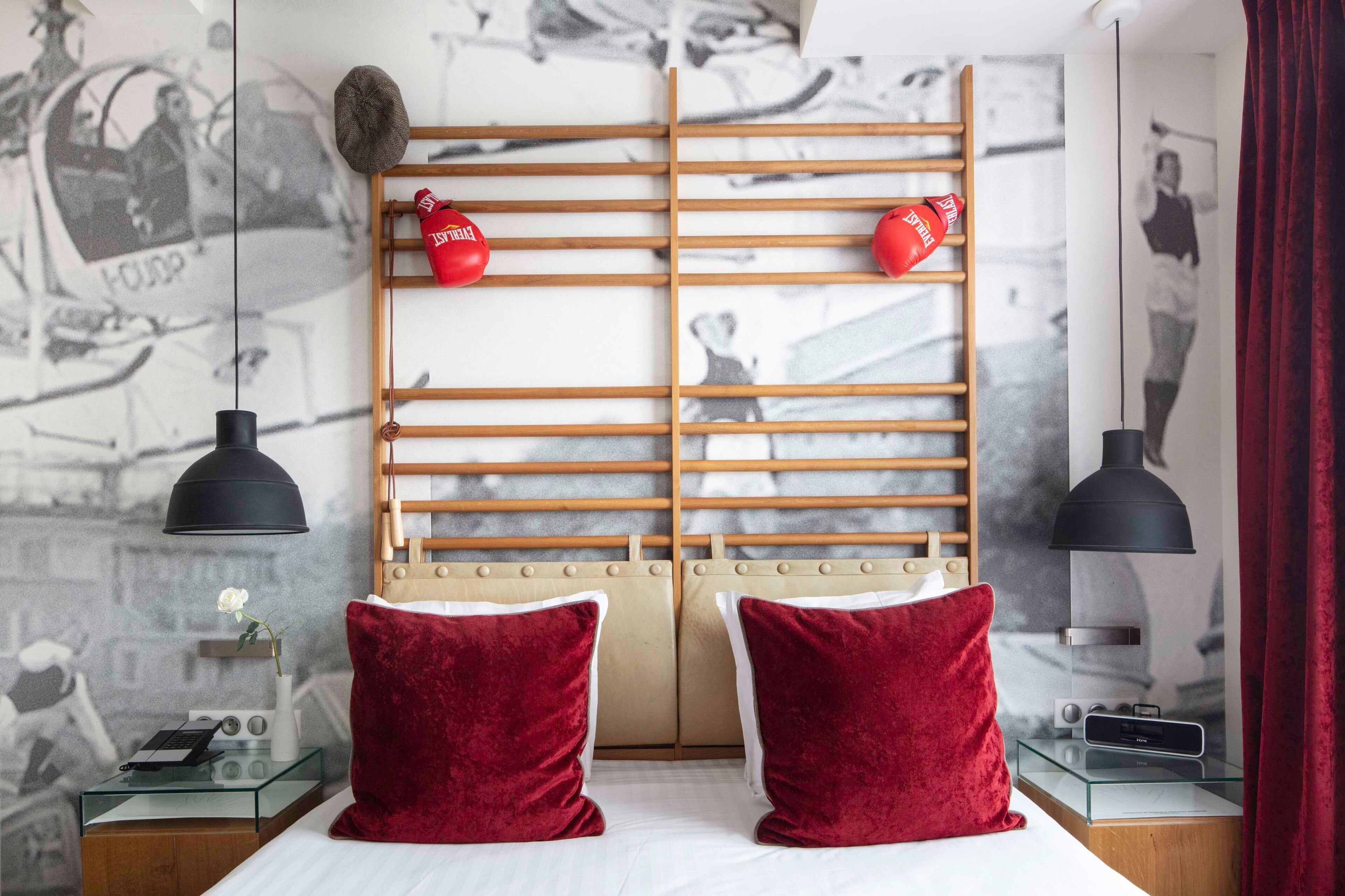 Habitación de hotel con una cama, dos almohadas rojas, un cabecero de madera, una mesita de vidrio con un radio y una pared con una fotografía en blanco y negro.