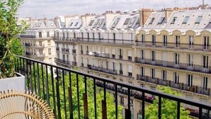 A balcony with a glass table and wicker chairs, overlooking a row of buildings with multiple stories and balconies.