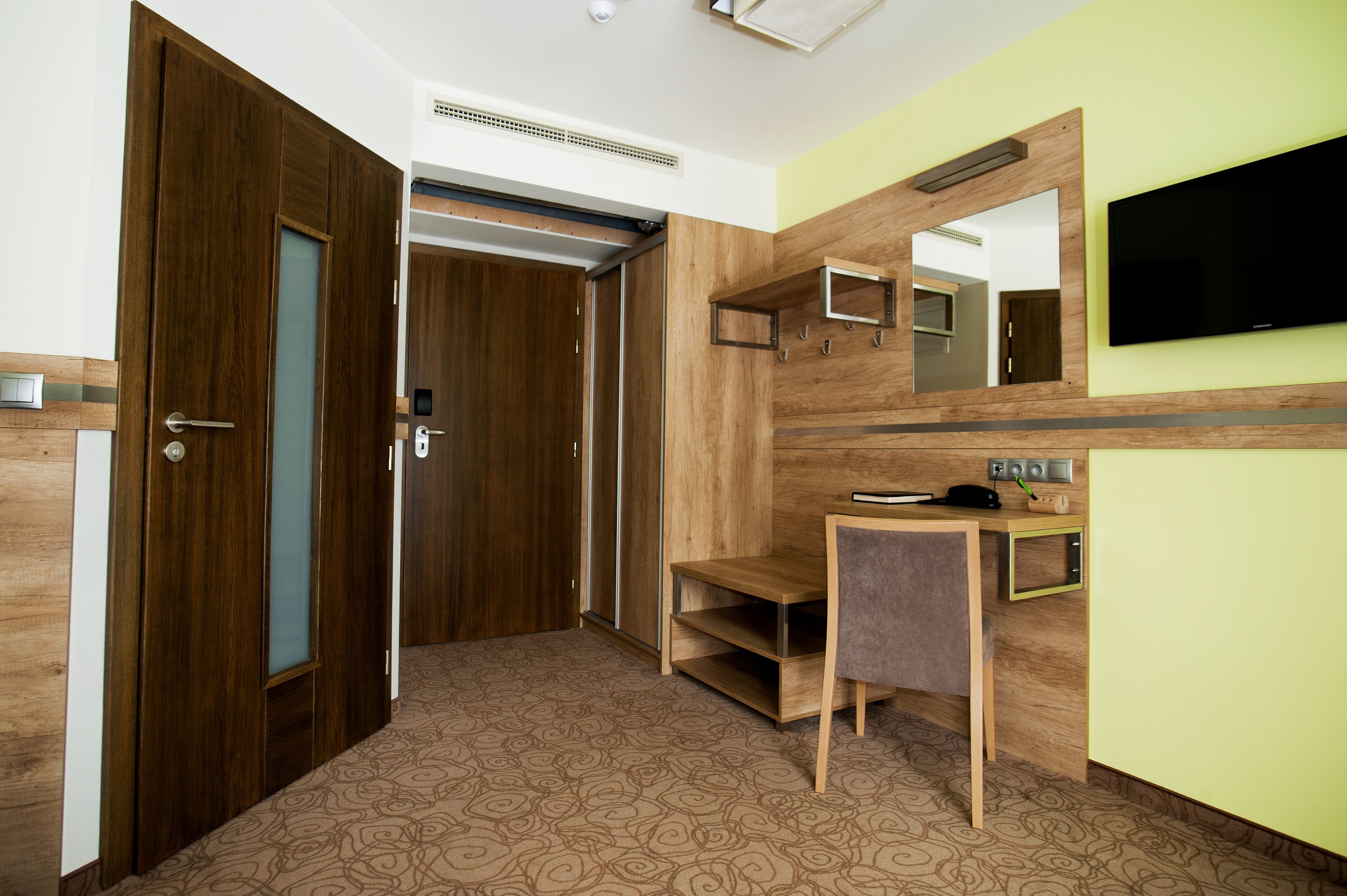 A hotel room with a wooden wardrobe, a desk with a chair, a television, and a mirror on the wall.