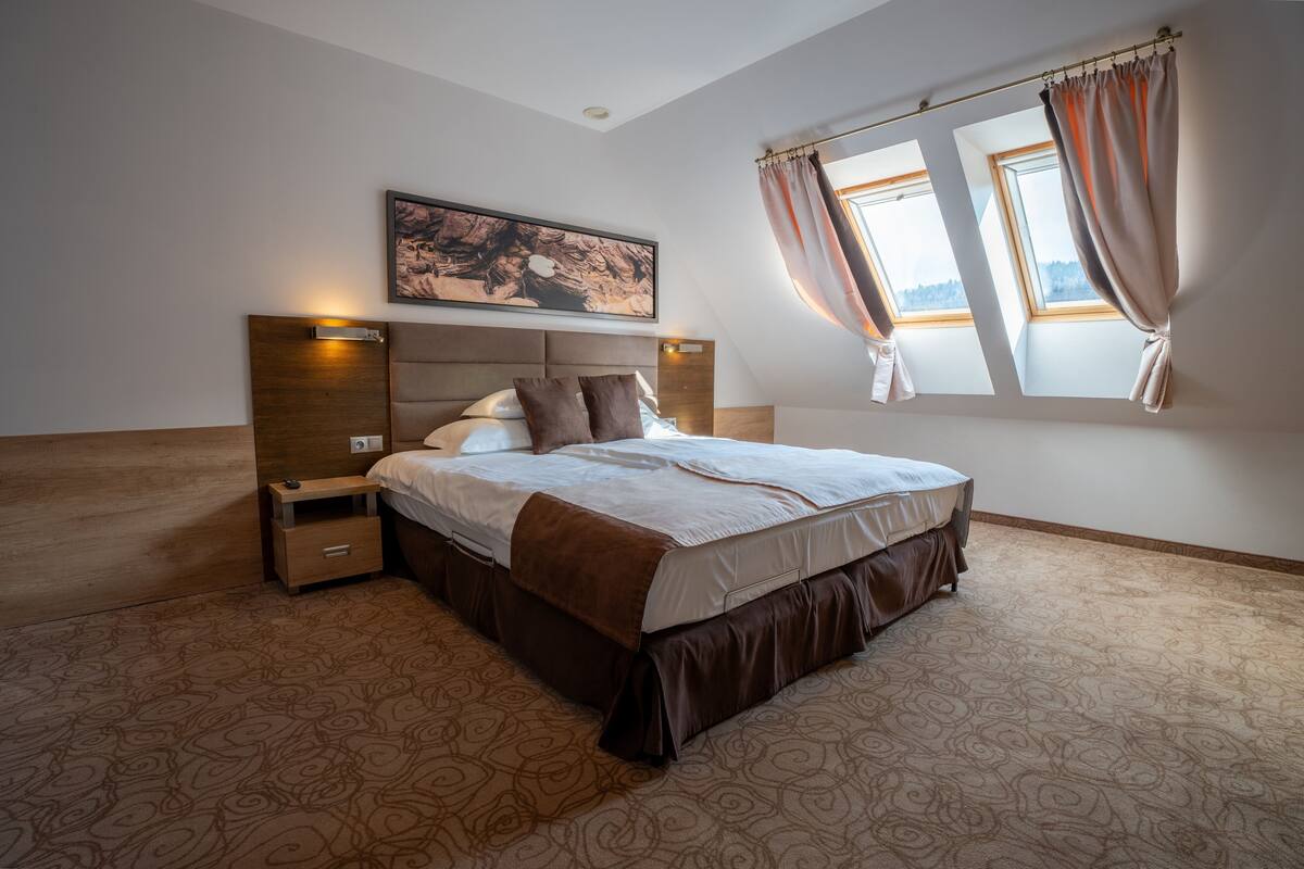 A bedroom with a large bed, a nightstand, a wall-mounted light, and a window with curtains.