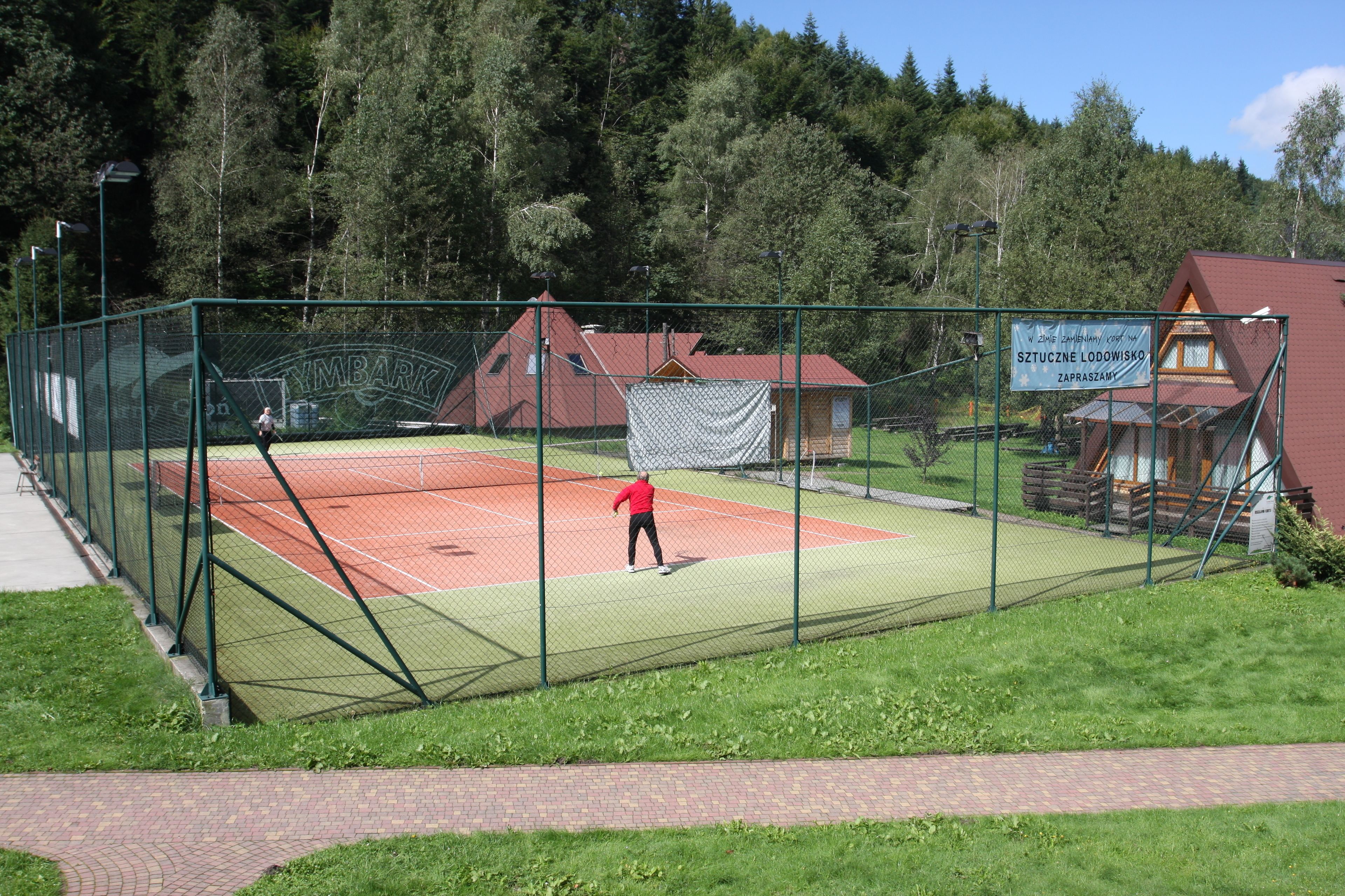 Una cancha de tenis con red, rodeada por una valla verde. Se observan edificios al fondo, uno con techo rojo.