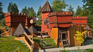 Un área de juegos con temática de castillo, con estructuras de madera, un puente colgante y un pequeño jardín.