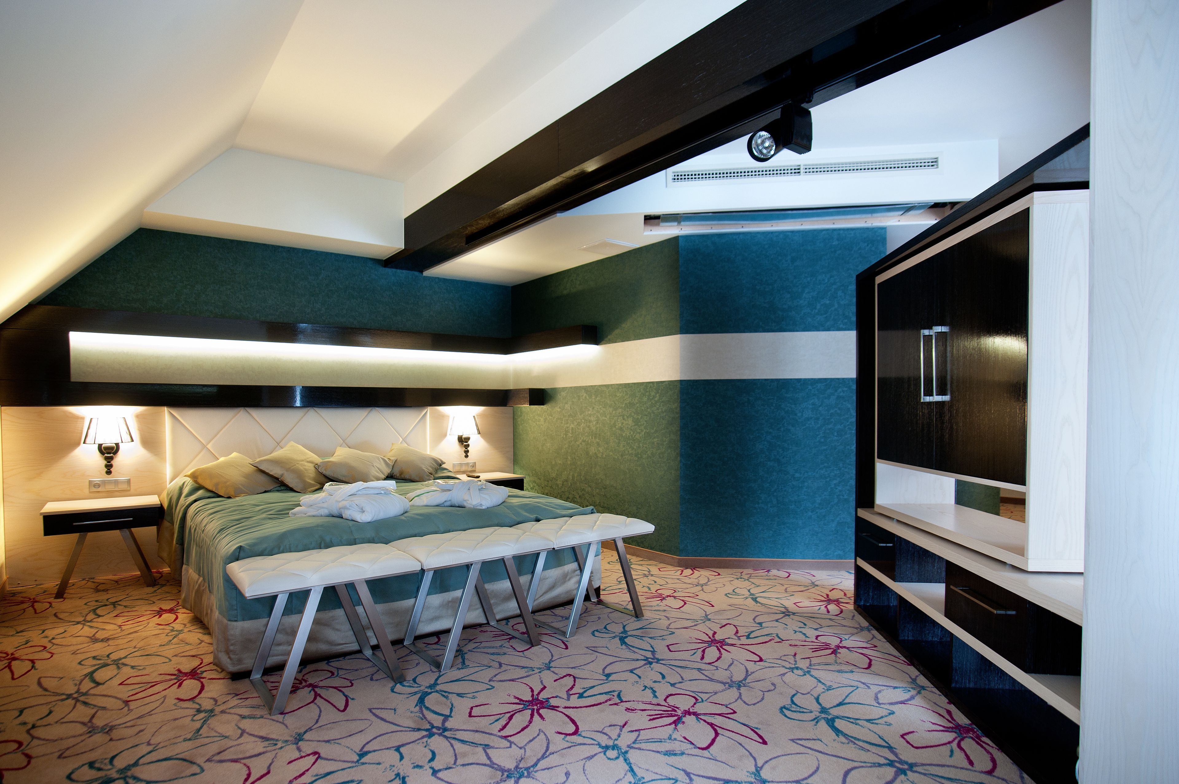 Habitación de hotel moderna con cama, banco, televisor y alfombra estampada.