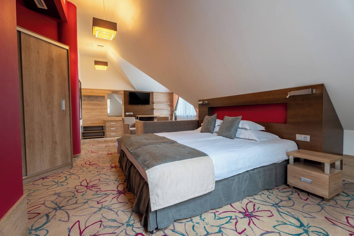 A modern hotel room with a large bed, a wooden wardrobe, a small bedside table, and a TV mounted on the wall.