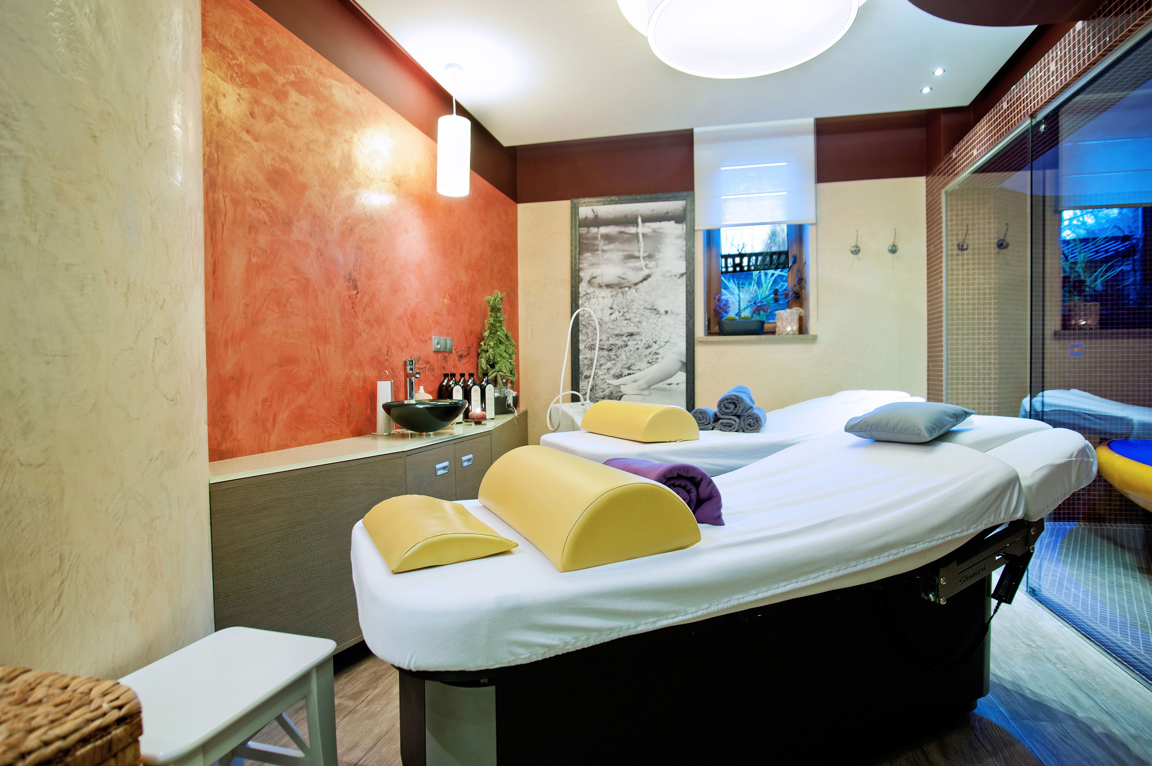 Une salle de spa moderne équipée de deux tables de massage, d’un petit lavabo et d’une fenêtre donnant sur l’extérieur.