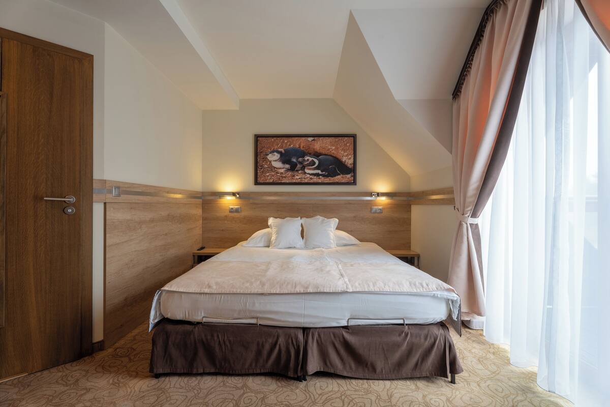 A bedroom with a large bed, wooden headboard, a painting above the bed, and a window with curtains.