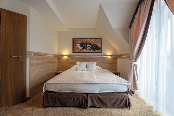 A bedroom with a large bed, wooden headboard, a painting above the bed, and a window with curtains. - Hotel & SPA Czarny Groń (Andrychow)