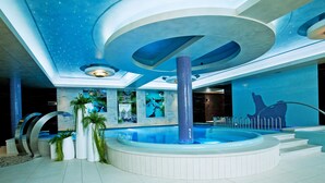 Un área de piscina interior moderna con un jacuzzi central, rodeado de asientos y plantas decorativas.