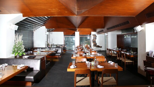 Ein geräumiges Restaurant mit hohen Decken und modernen Holzmöbeln. Die Tische sind ordentlich mit Stühlen bestückt, und Pflanzen sorgen für einen Hauch von Grün.