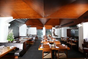 A spacious restaurant with high ceilings and modern wooden furniture. Tables are neatly arranged with chairs, and there are plants adding a touch of greenery. - Hotel Royal Garden (Mumbai)