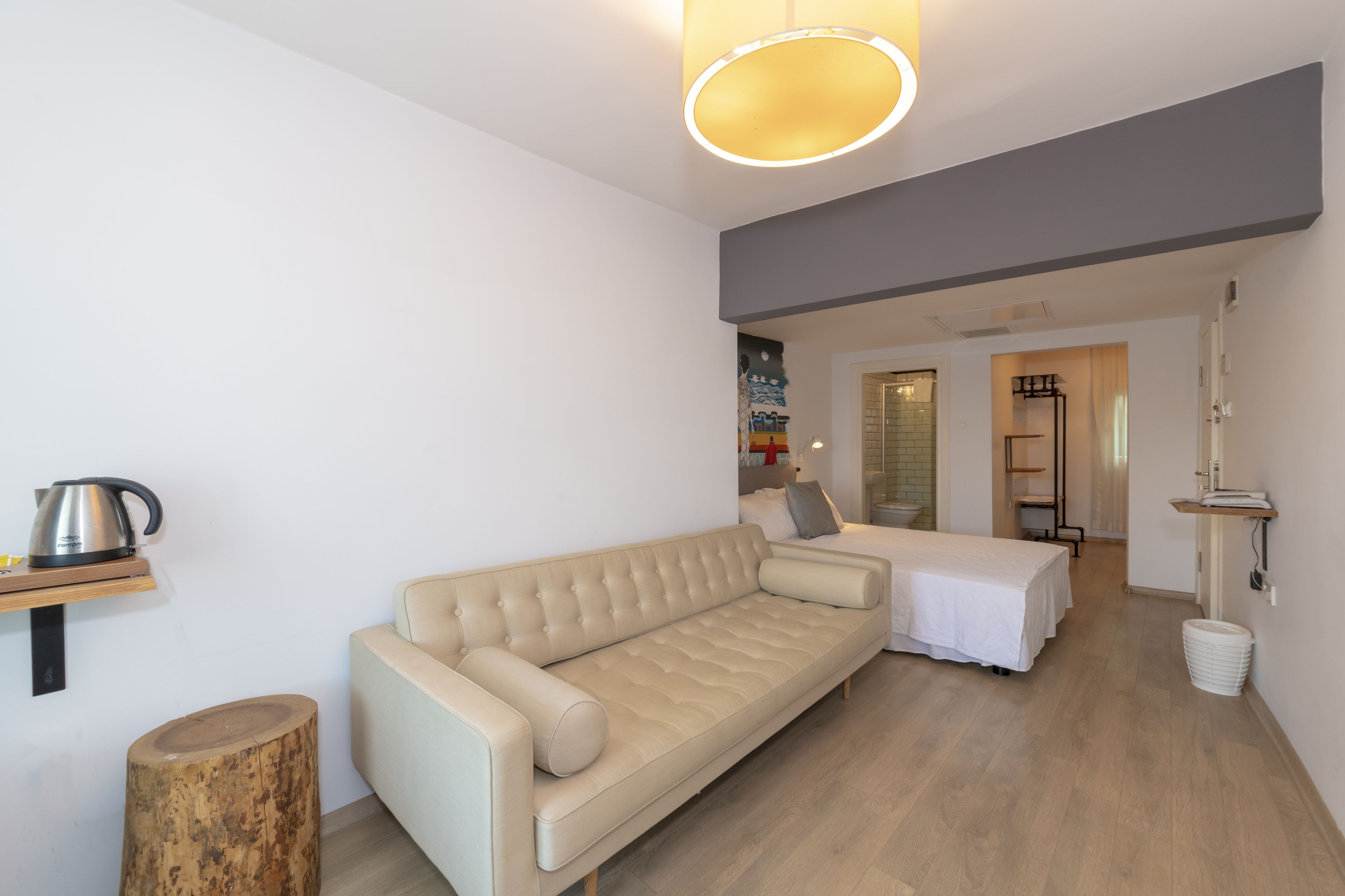 Une pièce moderne et bien éclairée, avec un canapé beige, une petite table sur laquelle se trouve une bouilloire, et un tabouret en bois. On y trouve un lit recouvert d’une courtepointe blanche et une table de chevet avec une lampe. La pièce est dotée d’un plancher en bois et de murs blancs.