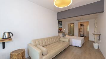 Une pièce moderne et bien éclairée, avec un canapé beige, une petite table sur laquelle se trouve une bouilloire, et un tabouret en bois. On y trouve un lit recouvert d’une courtepointe blanche et une table de chevet avec une lampe. La pièce est dotée d’un plancher en bois et de murs blancs.