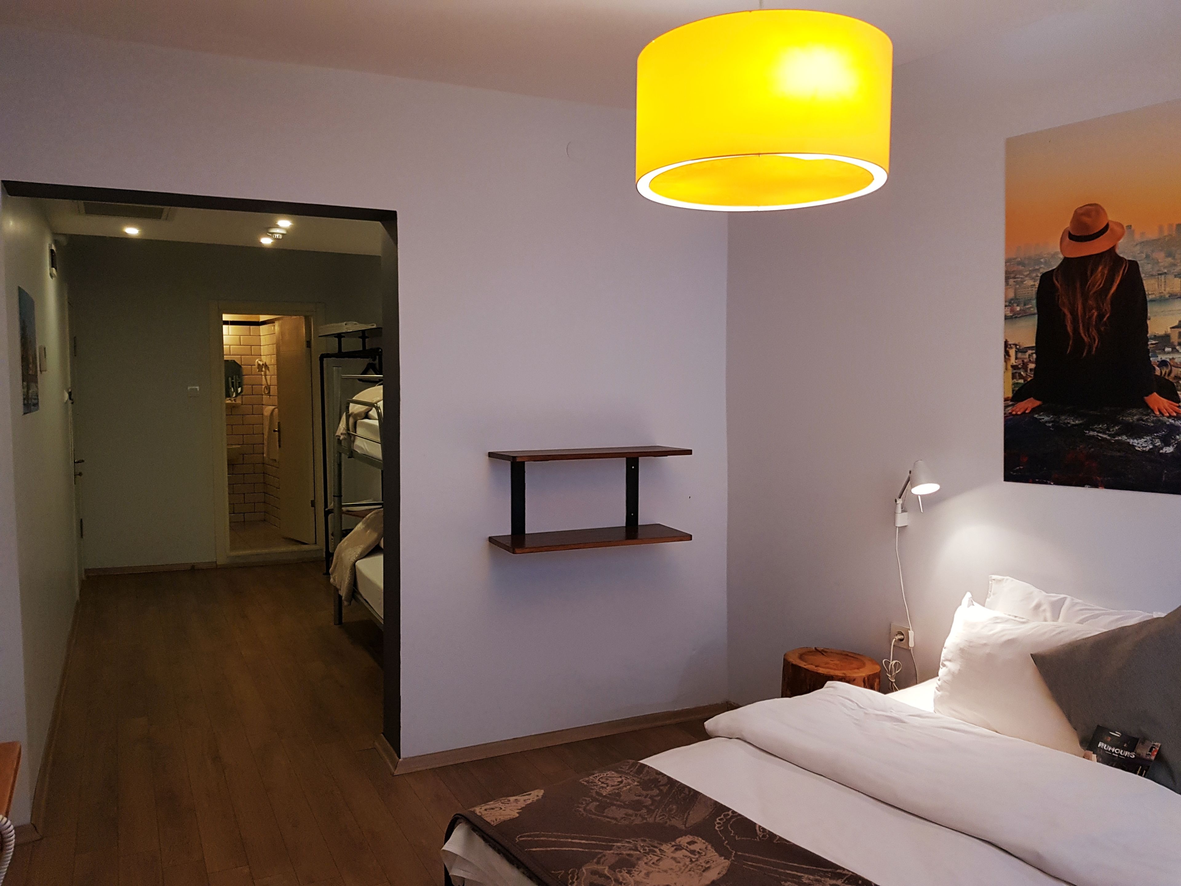 A hotel room with a bed, a bedside table, a hanging light fixture, a wall-mounted shelf, and a poster.