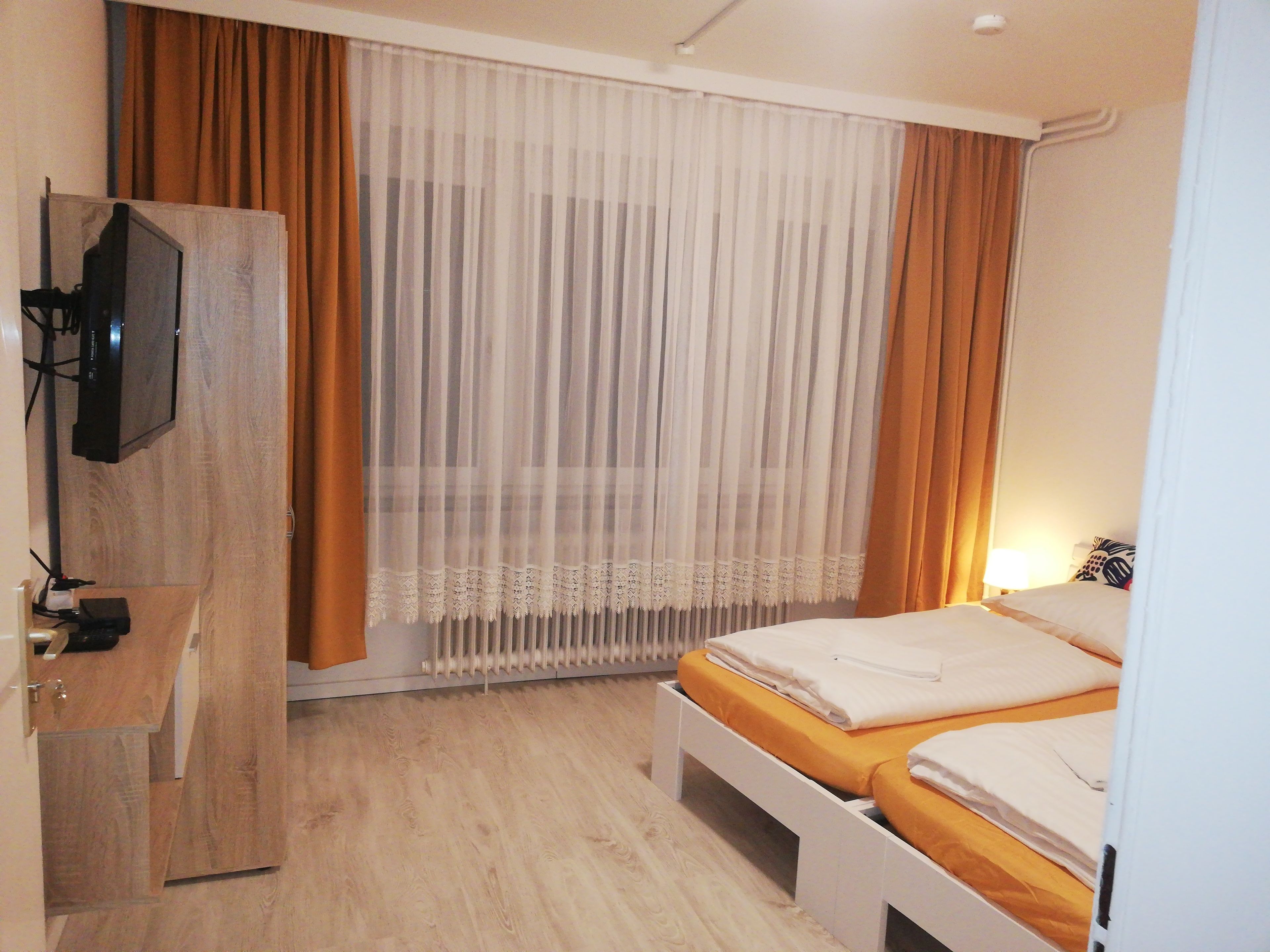 Ein Schlafzimmer mit einem Bett, einem an einer Holzkommode montierten Fernseher und einem Fenster mit durchsichtigen Vorhängen.