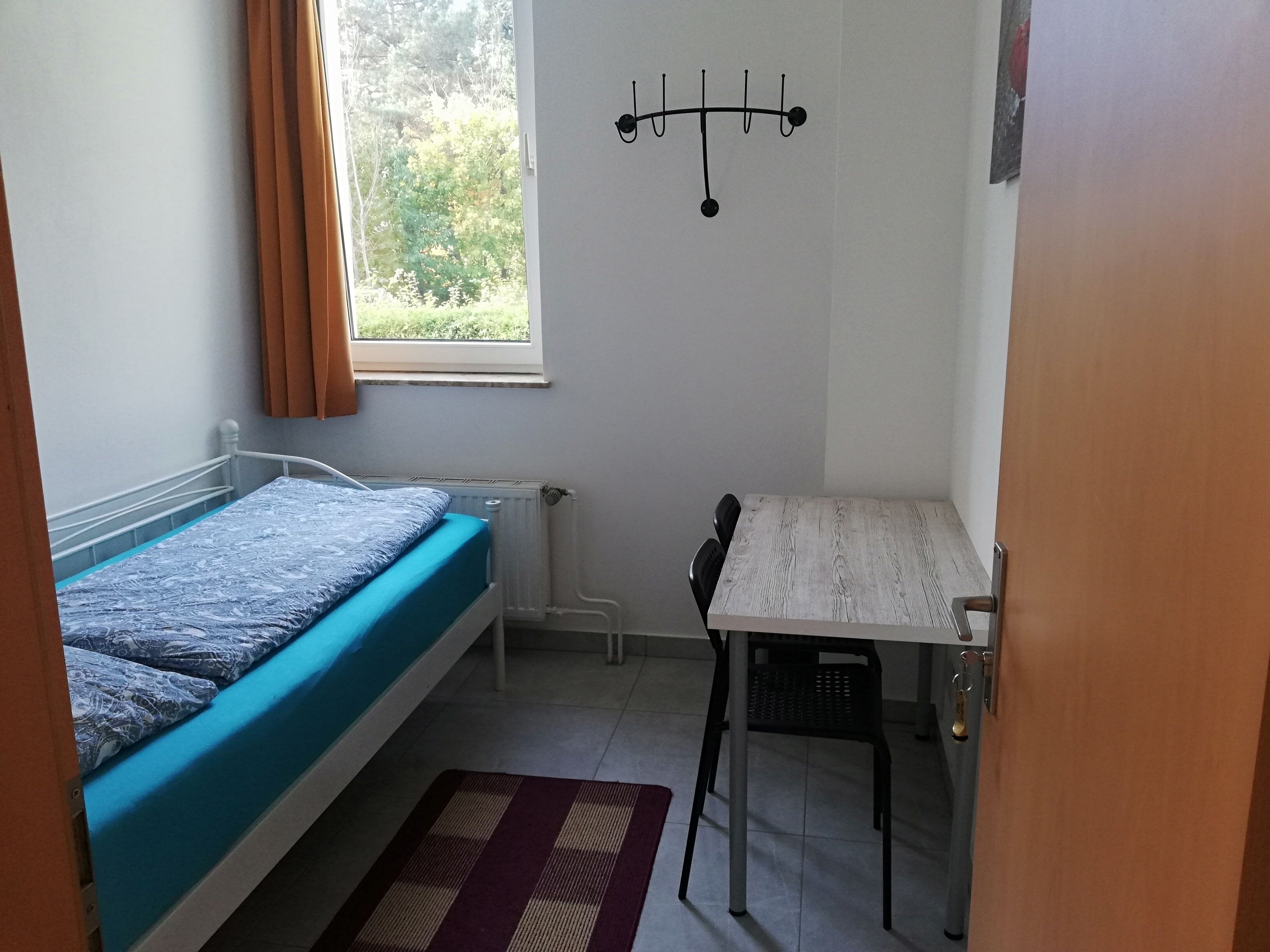 Una habitación pequeña y ordenada con una cama individual, una mesa y una silla. Se ve una ventana que proporciona luz natural y un radiador debajo de la cama.