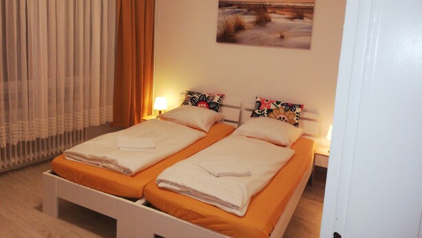 A hotel room with two single beds, a painting of a sunset, a window with curtains, and a nightstand with a lamp. - The Hostel (Hamburg)