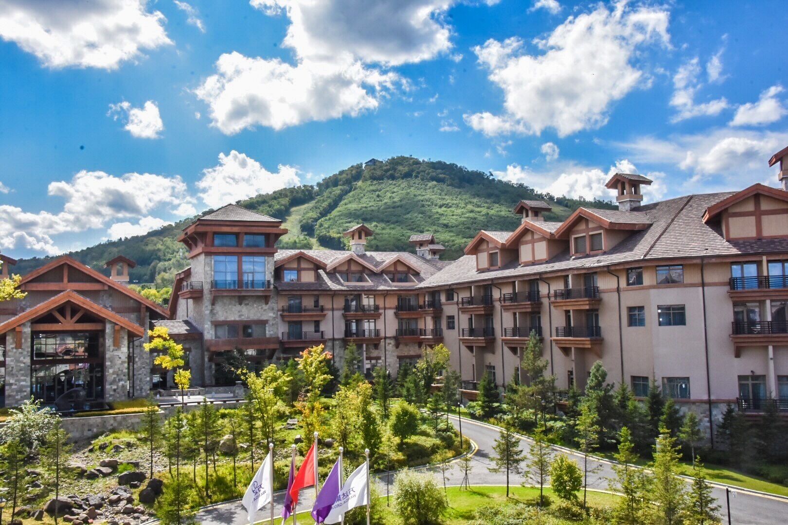 A multi-story hotel with a mountain backdrop, featuring balconies, a stone facade, and a landscaped courtyard.