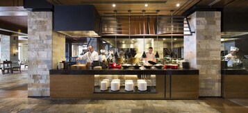 Una cocina moderna con chefs preparando comida, pilares de piedra y una barra con platos apilados.